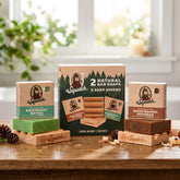 Dr. Squatch Wood Barrel Bourbon and Rainforest Rapids bar soaps displayed on cedarwood soap savers beside their cardboard boxes and kit packaging.