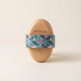 beechwood dry body brush standing upright with tropical flamingo-patterned cotton strap wrapped around middle against neutral beige background