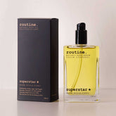 routine Superstar Botanic Body Oil in 100ml glass bottle with black pump next to charcoal-black cardboard packaging showing warm, rich & iconic scent description