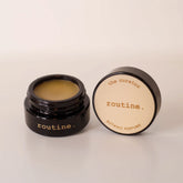 Routine The Curator botanic perfume balm - open black glass jar showing golden-yellow solid perfume with cream-coloured lid displaying product name
