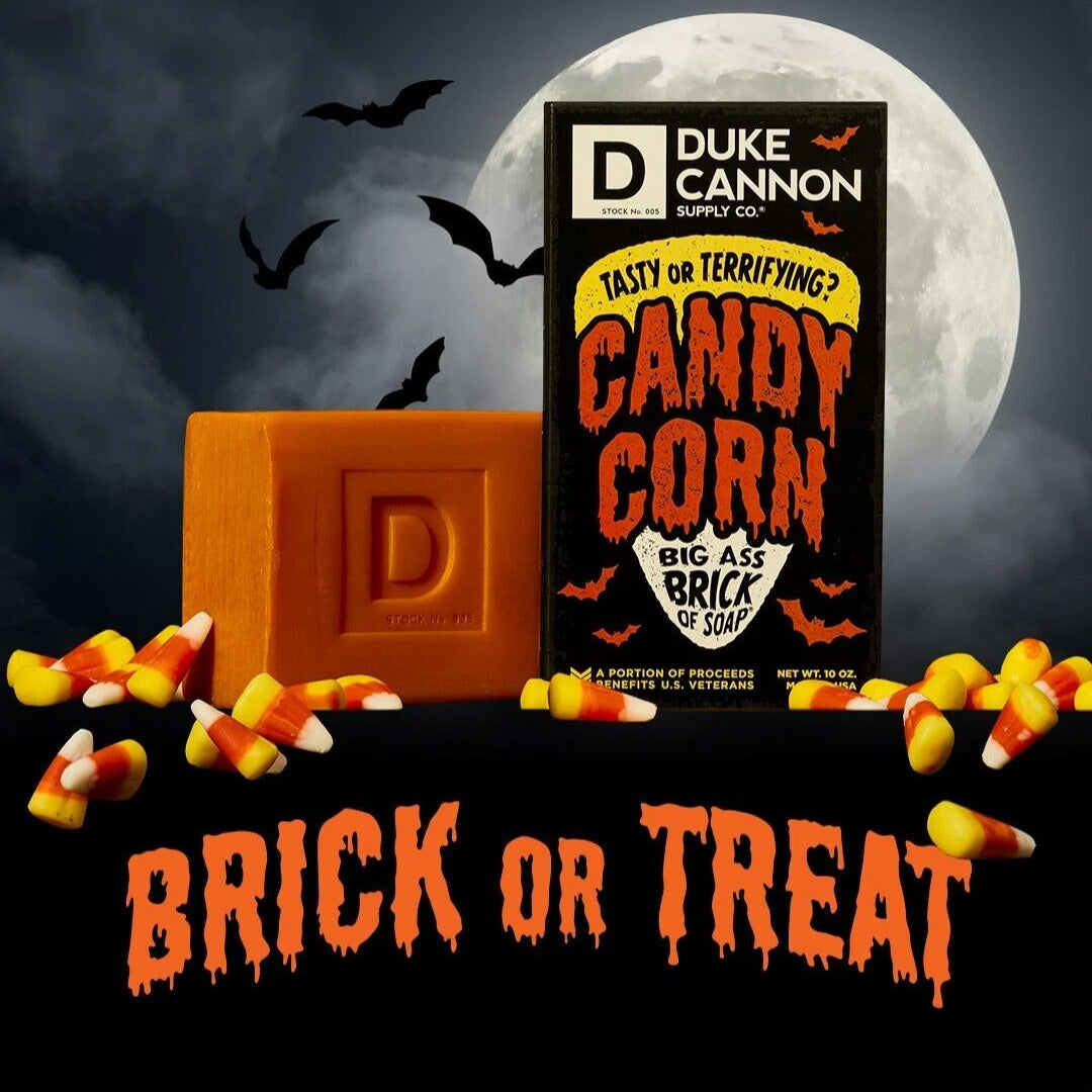 Halloween-themed Duke Cannon Candy Corn soap and black box surrounded by candy corn pieces against spooky nighttime backdrop with full moon and flying bats