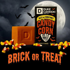 Halloween-themed Duke Cannon Candy Corn soap and black box surrounded by candy corn pieces against spooky nighttime backdrop with full moon and flying bats