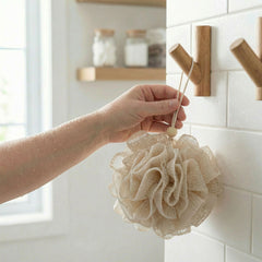 Hand holding a beige loofah against a tiled bathroom wall with wooden hooks.