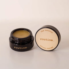 Routine The Curator botanic perfume balm - open black glass jar showing golden-yellow solid perfume with cream-coloured lid displaying product name