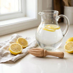 Beechwood lemon squeezer on a kitchen counter beside halved lemons and a glass pitcher of lemon water