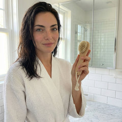 Woman in white bathrobe holding wooden dry brush with boar bristles in bright bathroom, demonstrating daily wellness self-care routine